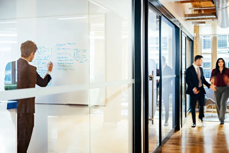 A colleague writes on a whiteboard as two others walk along a glass-walled hallway in the office.