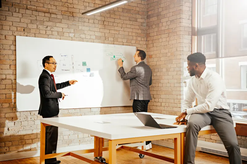 Three colleagues work through a plan at a whiteboard in a brick-walled meeting room.