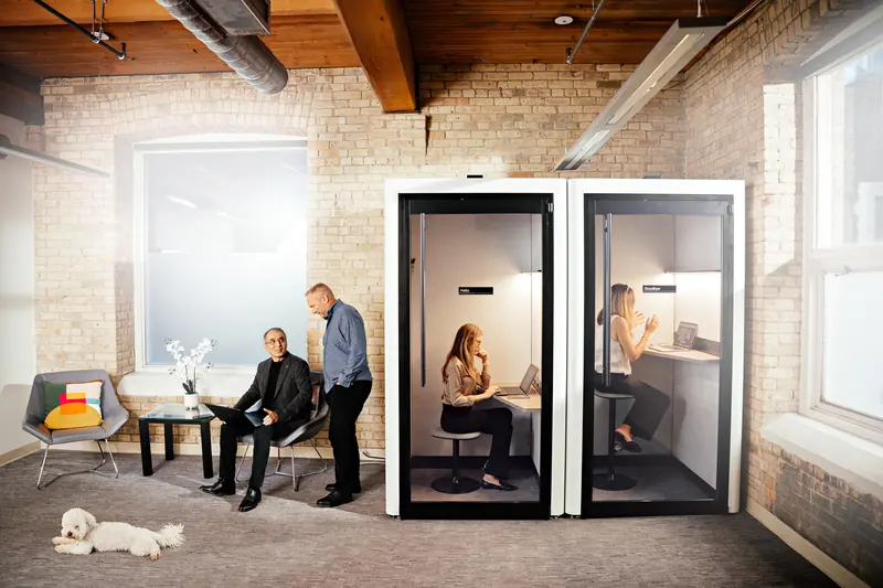 Colleagues talk in the office lounge beside two phone booths, with a dog asleep on the floor.